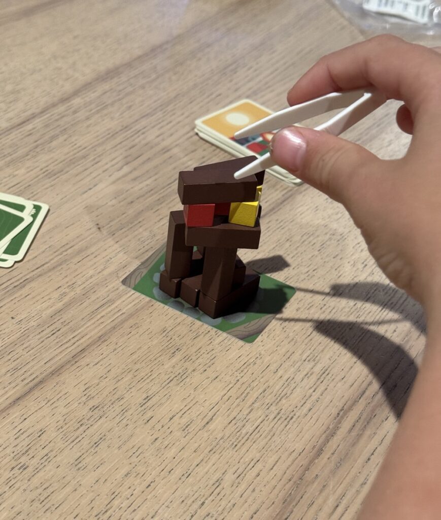 A child's hand holding tweezers after placing a block on a pile of other blocks