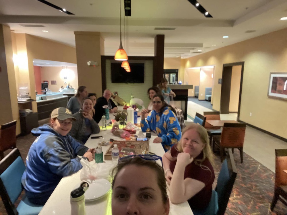 Friends gathered around a dinner table in Arkansas, smiling at the camera with food and drinks including Tim Tams and Vegemite